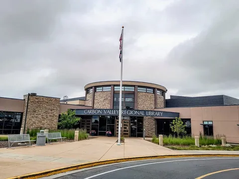 The Carbon Valley Regional Library on Park Avenue in Firestone under a grey summer sky: a round stone-and-glass entrance tower behind a flagpole, with the library's name in white letters over the doors and benches on the walk.