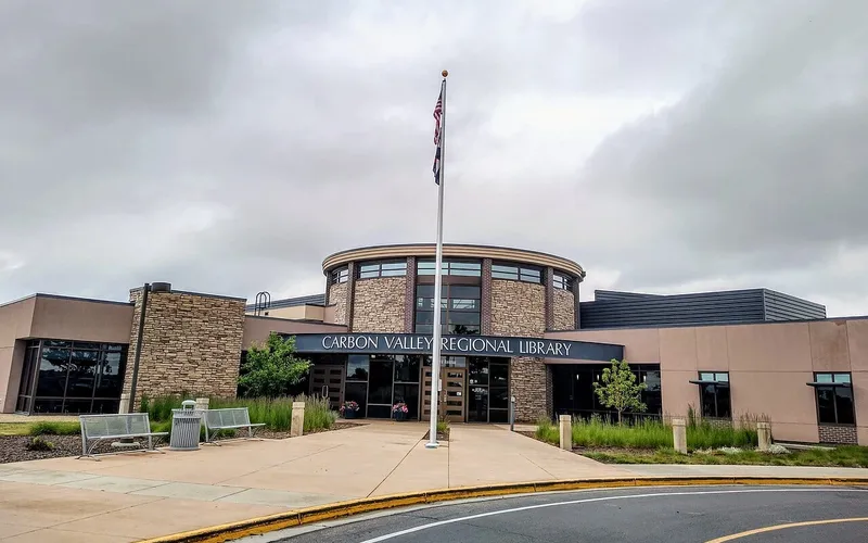 The Carbon Valley Regional Library on Park Avenue in Firestone under a grey summer sky: a round stone-and-glass entrance tower behind a flagpole, with the library's name in white letters over the doors and benches on the walk.