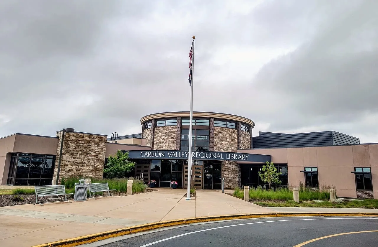 The Carbon Valley Regional Library on Park Avenue in Firestone under a grey summer sky: a round stone-and-glass entrance tower behind a flagpole, with the library's name in white letters over the doors and benches on the walk.