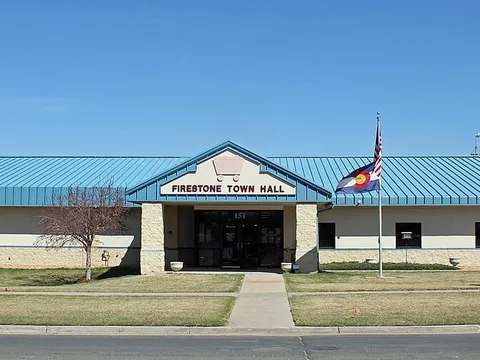 Firestone's former Town Hall at 151 Grant Avenue in the original townsite in March 2015: a low beige building with a blue metal roof and a gable reading Firestone Town Hall, with the Colorado and US flags out front under a clear sky.