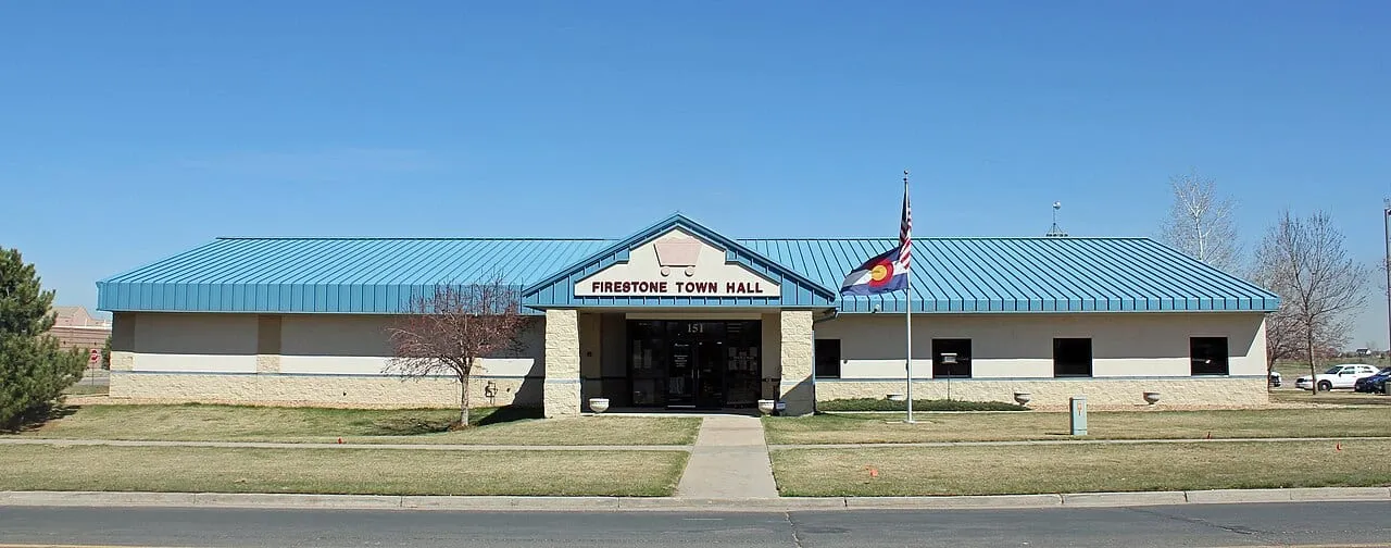 Firestone's former Town Hall at 151 Grant Avenue in the original townsite in March 2015: a low beige building with a blue metal roof and a gable reading Firestone Town Hall, with the Colorado and US flags out front under a clear sky.