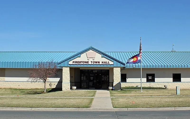 Firestone's former Town Hall at 151 Grant Avenue in the original townsite in March 2015: a low beige building with a blue metal roof and a gable reading Firestone Town Hall, with the Colorado and US flags out front under a clear sky.